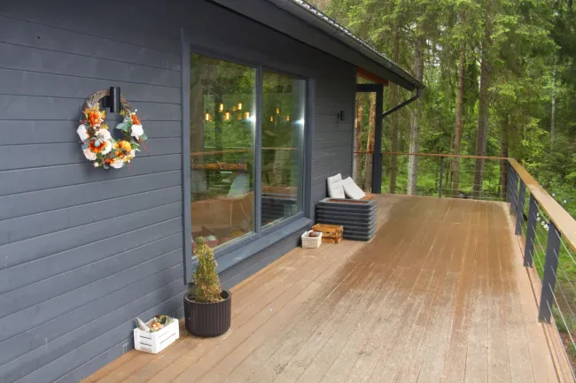 Exterior of a gray cabin with large window and wood deck overlooking a forest. A wreath hangs on the wall.