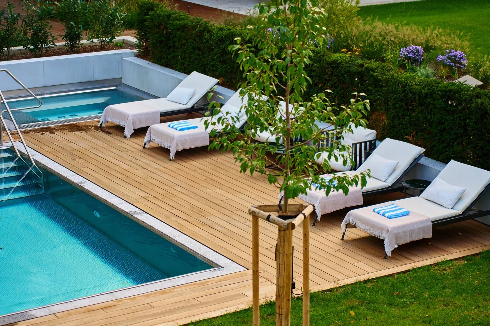 A swimming pool with lounge chairs on a wooden deck, next to a smaller hot tub and a green garden.