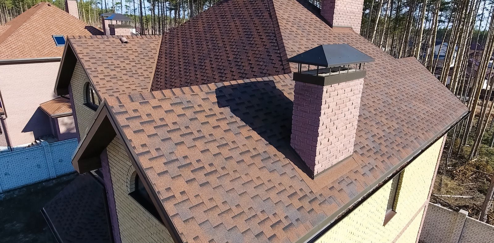 A high-angle view of a brown shingled residential roof with a brick chimney and a metal cap, set against a wooded background.