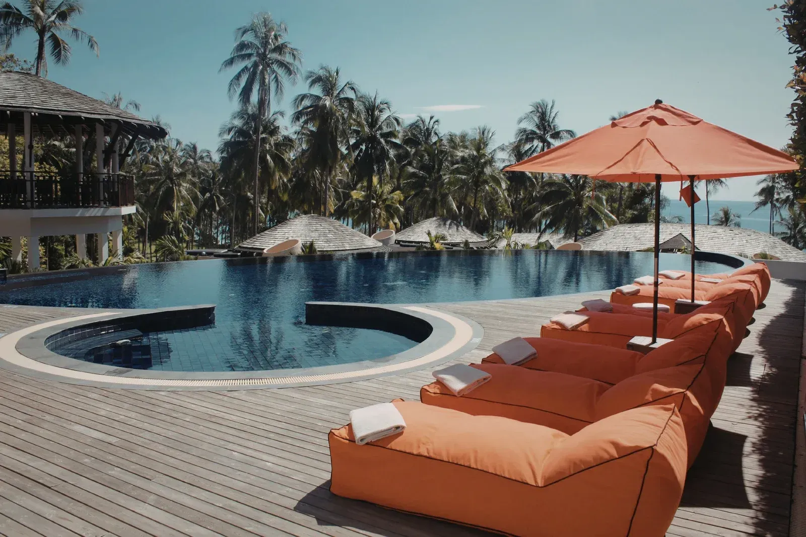 Orange lounge chairs and umbrellas arranged on a wooden deck next to a scenic swimming pool surrounded by palm trees.