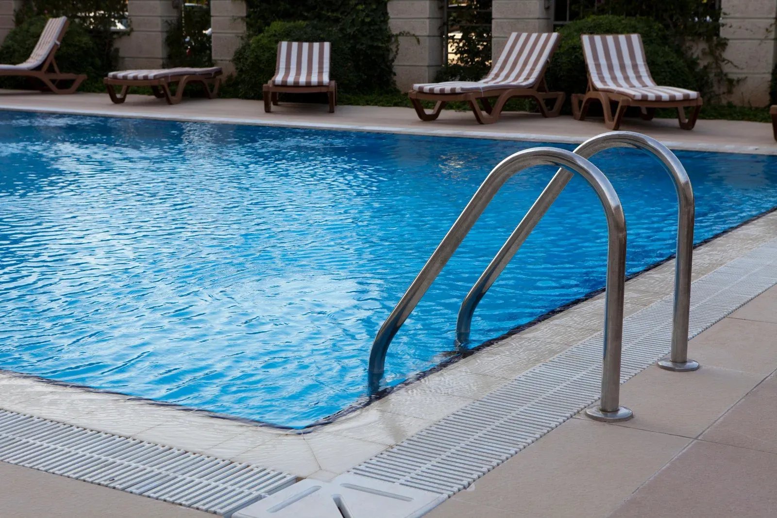 A rectangular blue swimming pool with metal steps in the foreground and a row of lounge chairs on the deck behind.