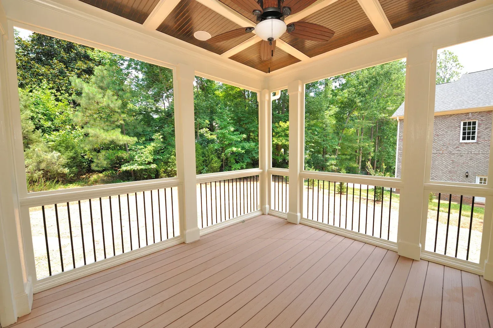 A covered wooden porch with a ceiling fan, white railings, and a view of trees and a brick house.