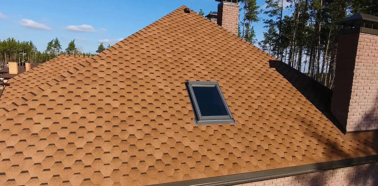 A brown hexagonal shingled roof with a rectangular skylight against a clear blue sky and trees.