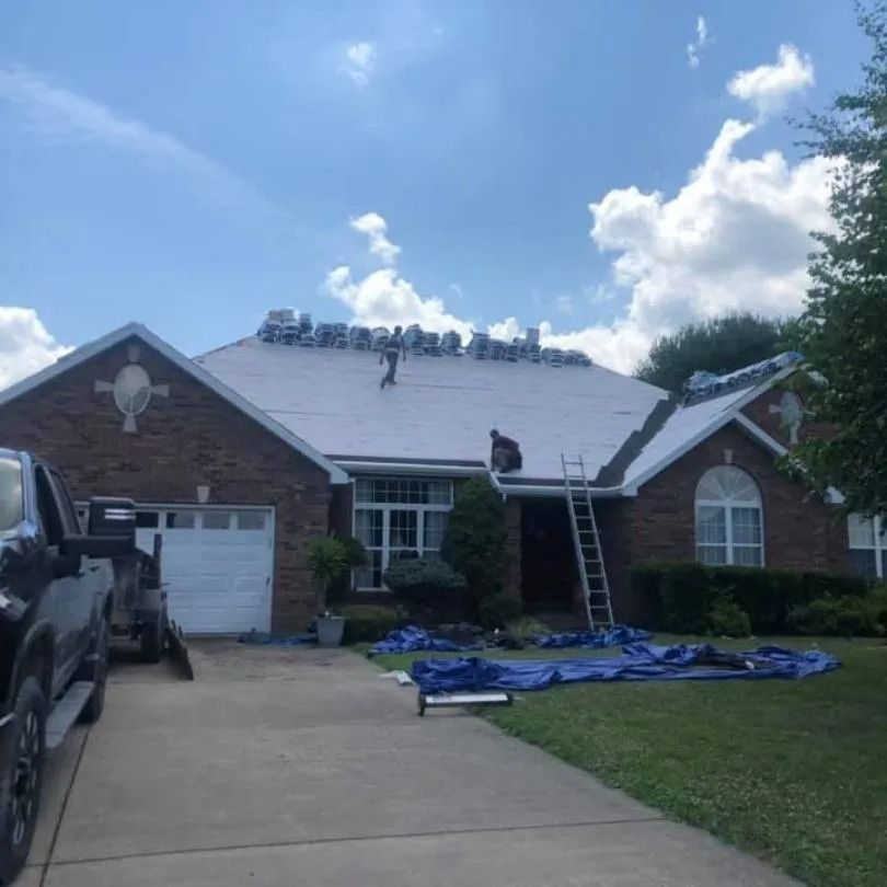 Workers installing a new roof on a brick house during the day, with materials on the roof and a ladder against the eaves.
