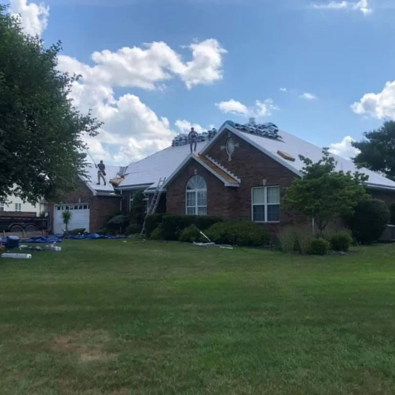 Workers perform roof repairs on a single-story brick house under a blue, cloudy sky.