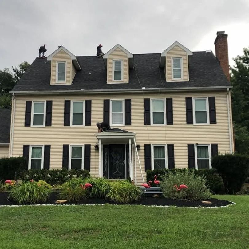 Workers repair the roof and front porch of a two-story beige house with black shutters and flamingo lawn ornaments.