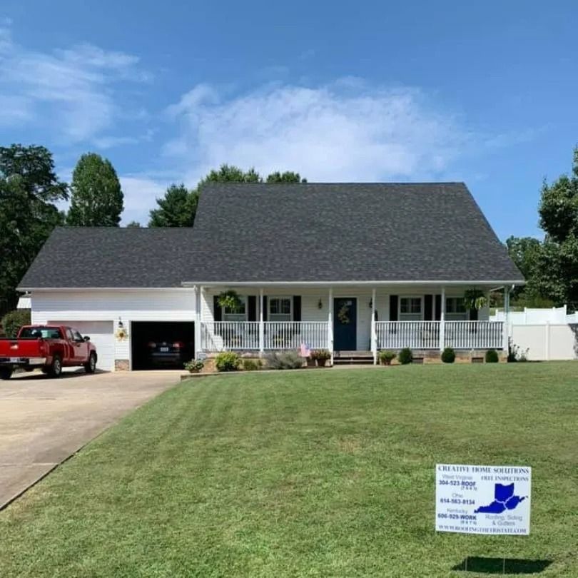 A white one-story house with a black roof, a front porch, and a garage, featuring a yard sign in front on a sunny day.