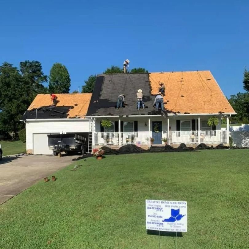 A crew works to install a new roof on a house under a clear blue sky, with a promotional sign in the front yard.