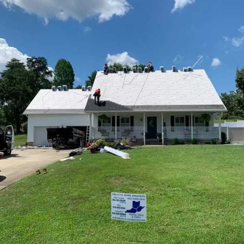 Workers install a new roof on a white residential house, with a company sign placed in the front lawn.