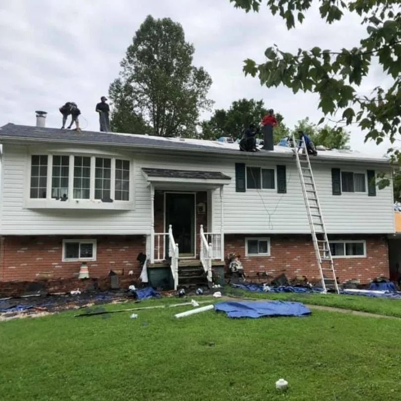 Workers repairing the roof of a two-story house with white siding and a brick base, with debris on the lawn below.
