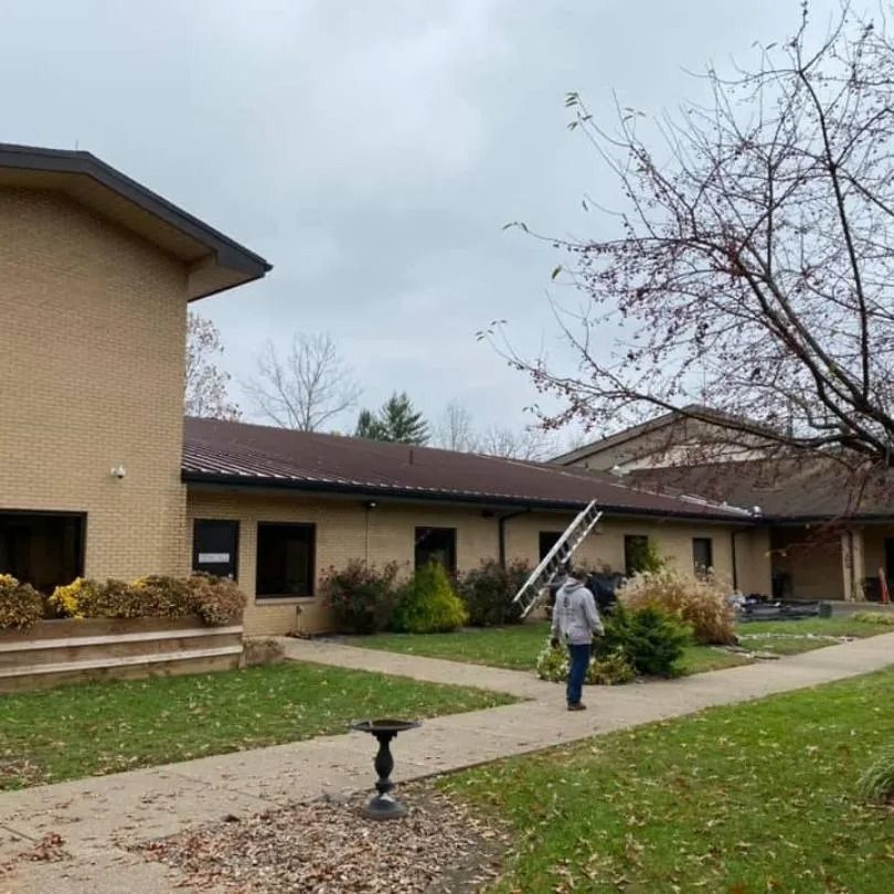 A person walks on a walkway toward a tan brick building, carrying a ladder over their shoulder on an overcast day.