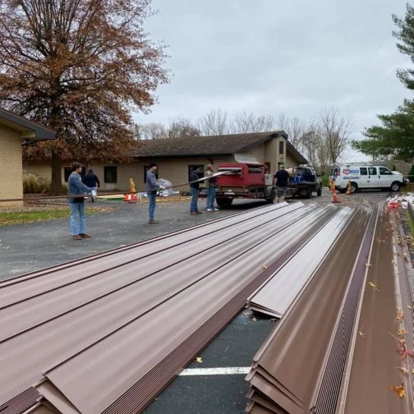 Workers use a red metal-forming machine to feed long, brown roofing panels onto a paved lot in front of a building.