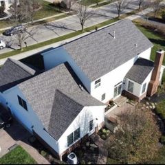 An aerial view of a two-story white residential house with a dark shingled roof, set next to a street and a grassy lawn.