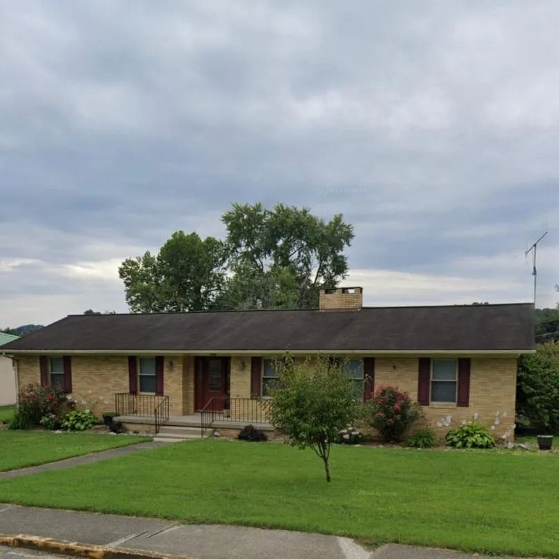 A one-story brick ranch-style house with a brown roof, dark shutters, and a front porch, surrounded by a green lawn.