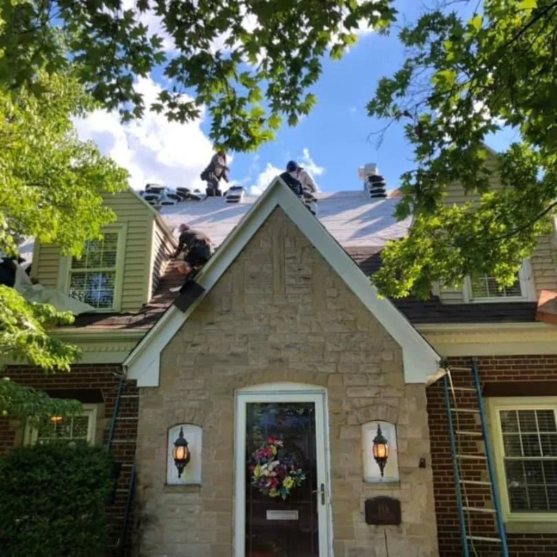 Workers on the roof of a stone and brick house during a roof replacement, viewed from ground level amidst green trees.