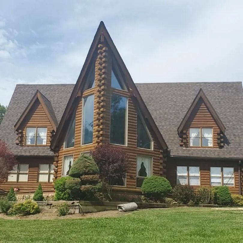A large, multi-story log cabin home with a prominent triangular front gable, peaked dormers, and a lush green lawn.