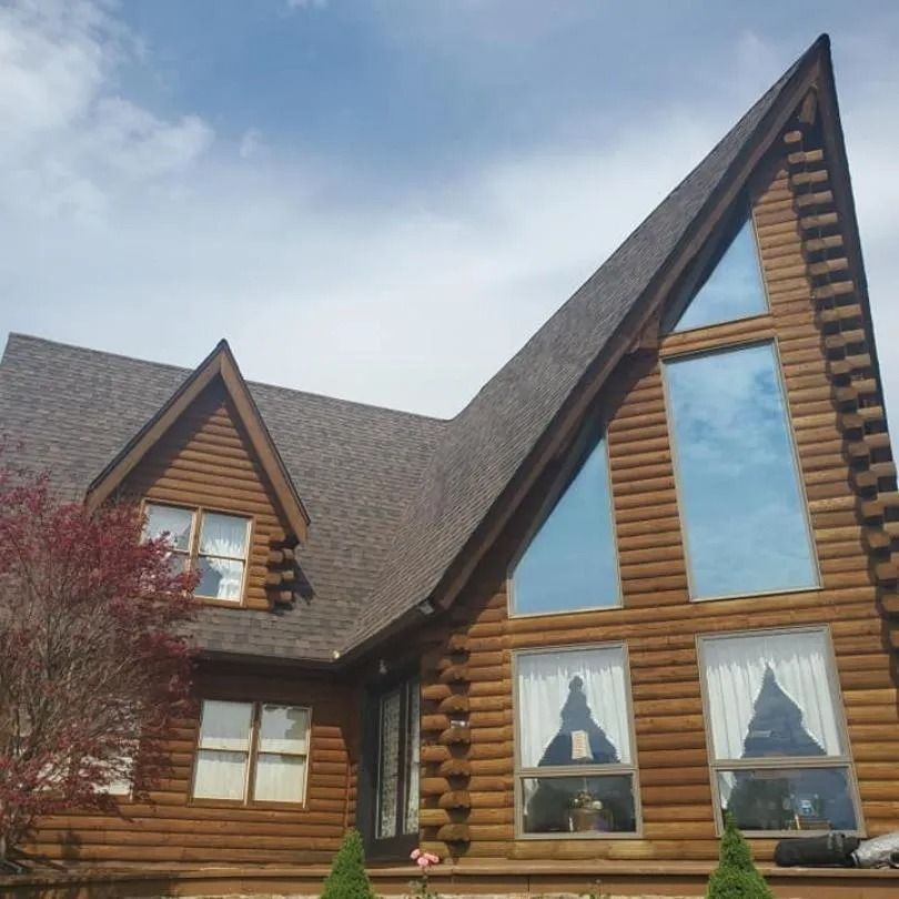 A two-story log cabin with a steep, angled roof and large windows set under a cloudy blue sky.