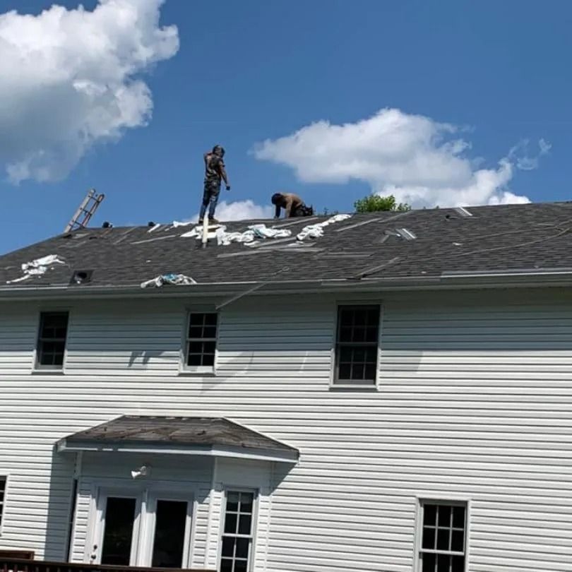 Two workers install shingles on the roof of a white, two-story house under a blue sky with scattered clouds.