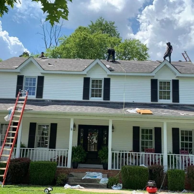 Workers replacing the shingles on the roof of a two-story white house with a front porch and dormer windows.