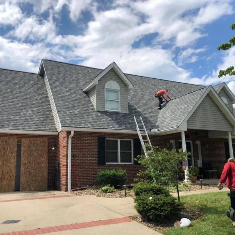 A worker in a red shirt walks on a gray shingled roof while a ladder leans against the side of a brick house.