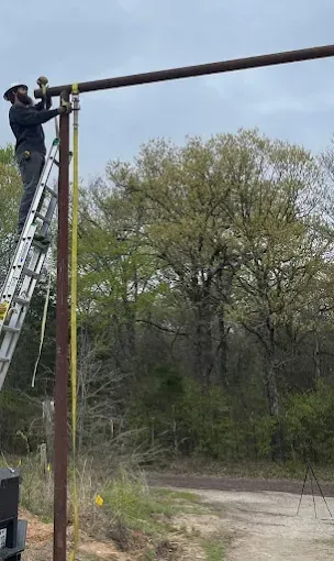 A person in a hard hat stands on a ladder, using a tool to attach a horizontal metal beam to a vertical pole outdoors.