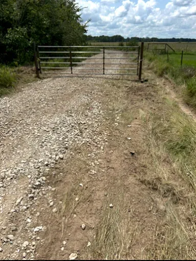 A metal farm gate blocks a gravel road leading into a rural field under a partly cloudy sky.