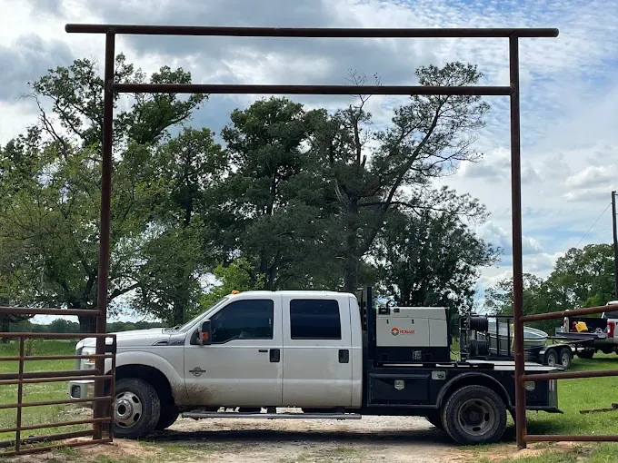 A white pickup truck with a large metal generator bed parked under a tall metal entrance gate on a rural lot.
