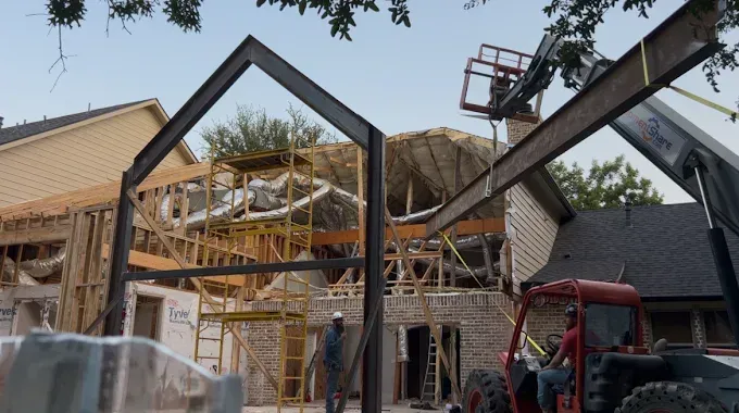 Construction site showing a large steel frame being positioned by a telehandler crane in front of a house under renovation.