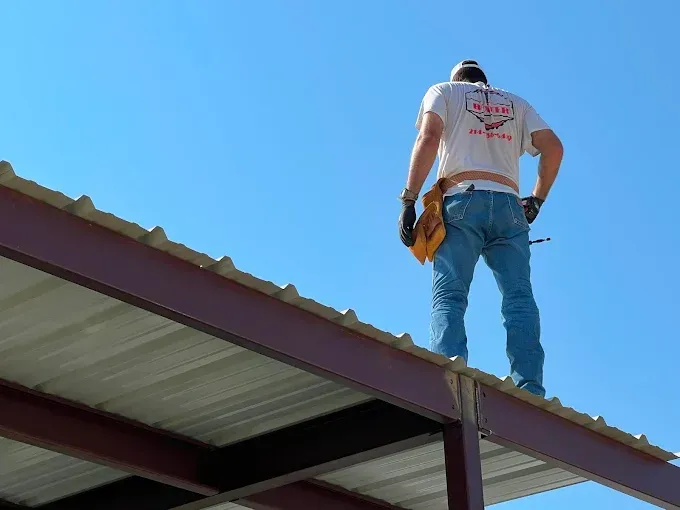A worker in a white t-shirt and jeans stands on the edge of a metal roof structure against a clear blue sky.