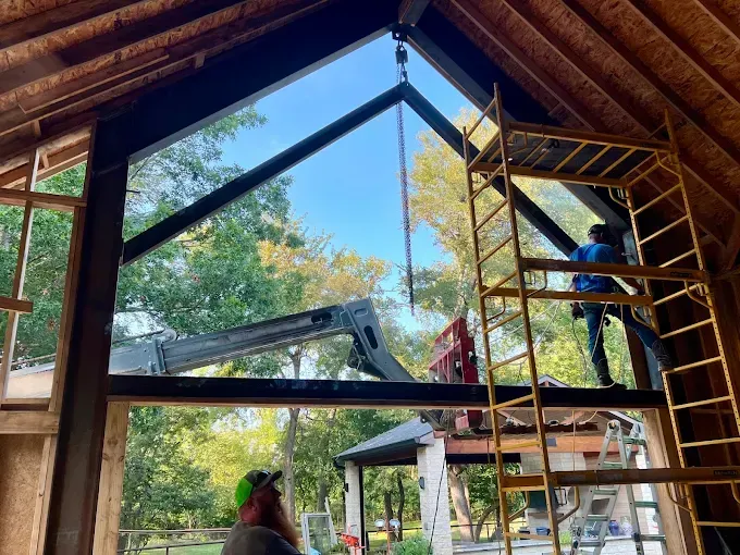 Construction workers install a large steel frame into the gable end of a building using a crane.
