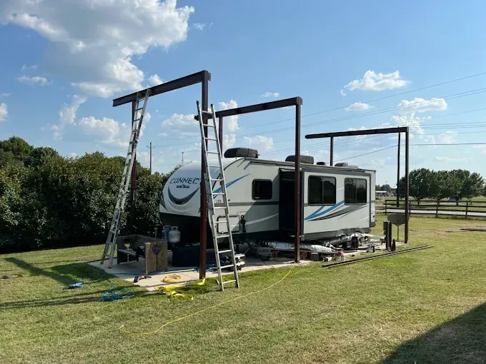 A travel trailer parked on a concrete pad under a series of tall, metal structural beams being installed by two ladders.