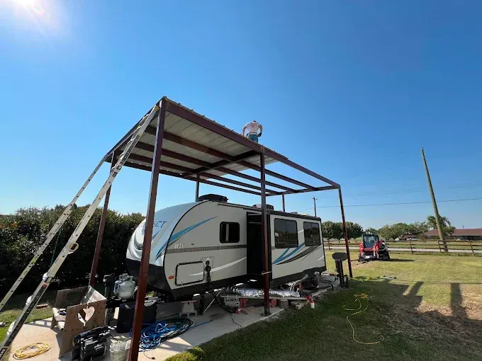 A person stands on top of a metal carport frame being built over a white and blue travel trailer in a grassy yard.