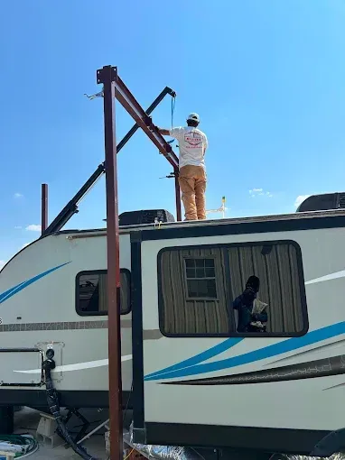 A person in work clothes stands on the roof of a camper, tethered to a metal frame structure outdoors under a blue sky.