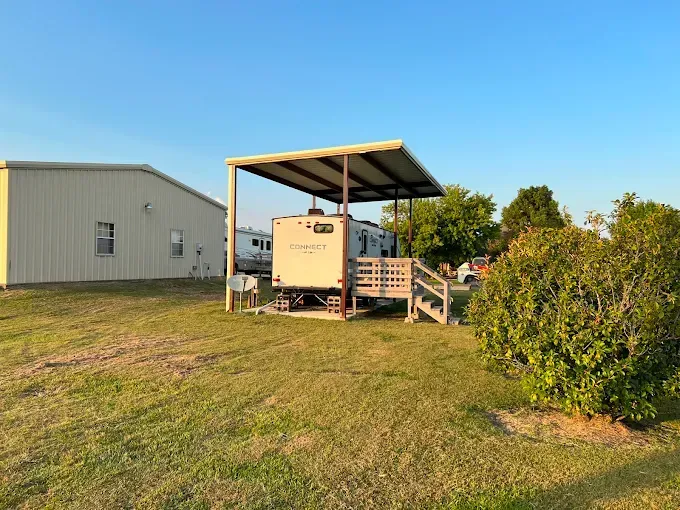 A camper trailer parked under a metal carport next to a white metal building in a grassy, sunlit yard.