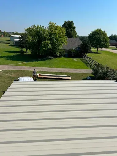 A view from a metal roof looking down at a person standing near long wooden boards on a grassy lawn by a house.