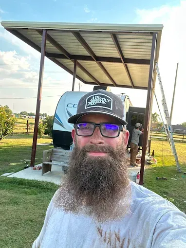 A man with a full beard and glasses poses for a selfie in front of a parked travel trailer under a metal carport.
