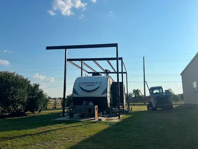 A travel trailer parked under a metal frame carport structure in a grassy yard on a sunny day with a skid steer nearby.