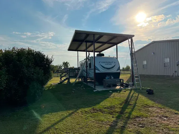 A white travel trailer parked under a metal carport structure in a grassy field on a sunny day.