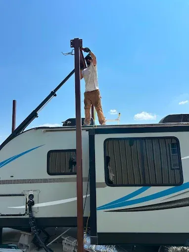 A person standing on the roof of a travel trailer, welding a tall, dark metal pole under a clear blue sky.