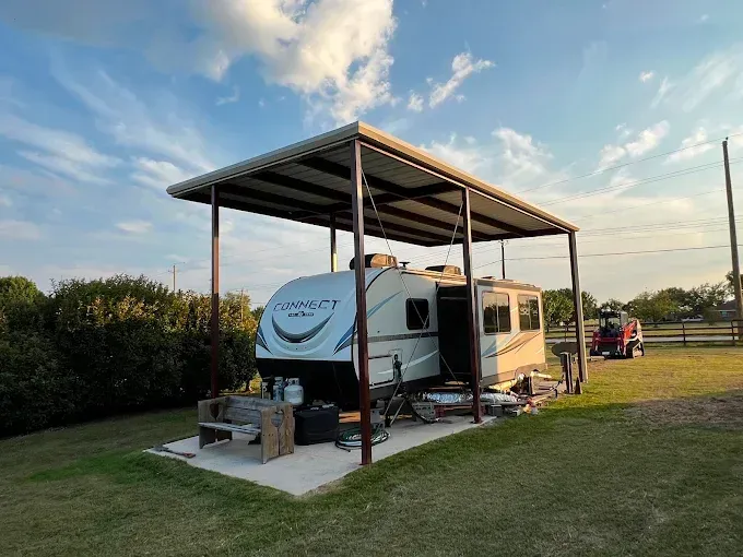 A white travel trailer parked under a metal carport structure on a concrete pad in a grassy field at sunset.