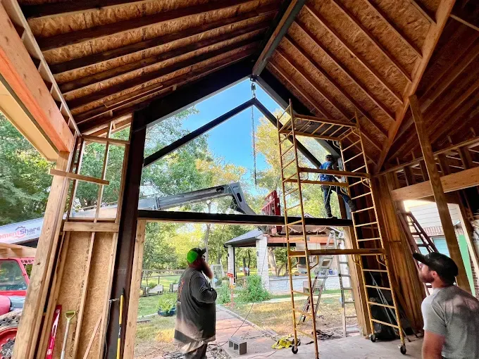 Construction workers assemble a large triangular window frame in the unfinished wood-framed wall of a house.