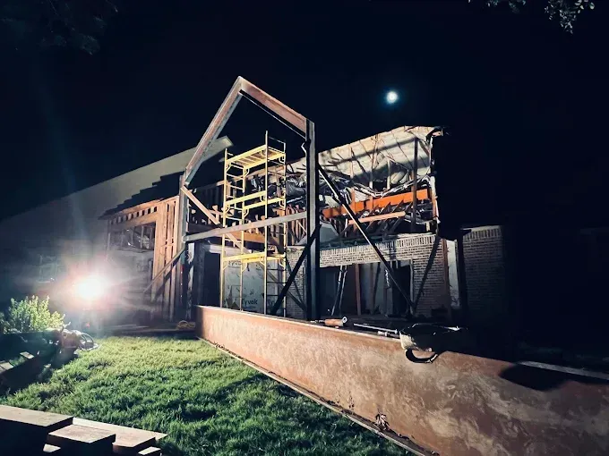 A steel frame for a building under construction at night, illuminated by a bright work light on a grassy lawn.