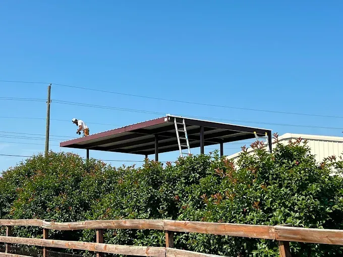 A person works on the metal roof of an open-sided carport structure outdoors, with dense green bushes and a wood fence.