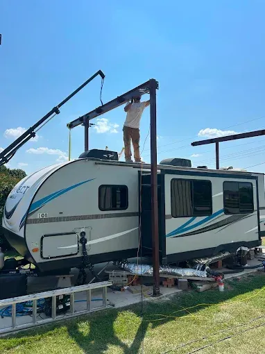 A person stands on the roof of a travel trailer under a metal frame structure outdoors on a sunny day.