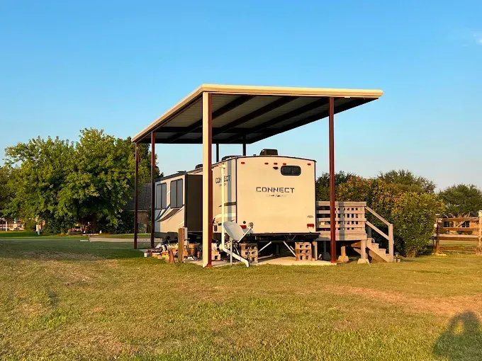 A white travel trailer parked under a metal carport structure in a grassy field on a clear, sunny day.