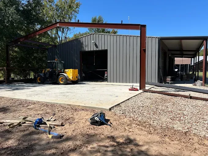 A construction site featuring a steel-framed workshop with gray metal siding and an attached carport under a blue sky.