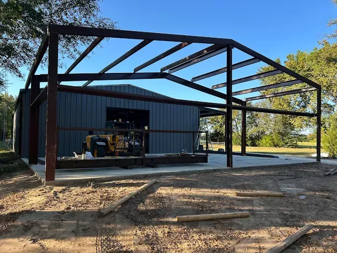 A metal building structure with an open porch frame addition attached to a closed workshop under a clear blue sky.