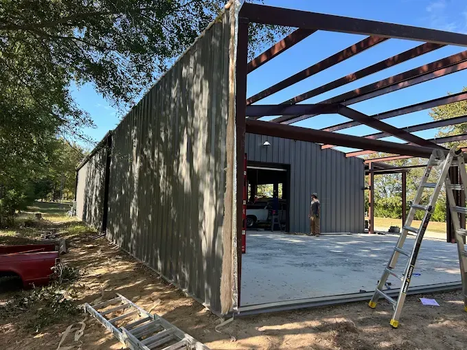 Construction of a metal building with an open roof frame, gray siding, and a concrete slab under a blue sky.