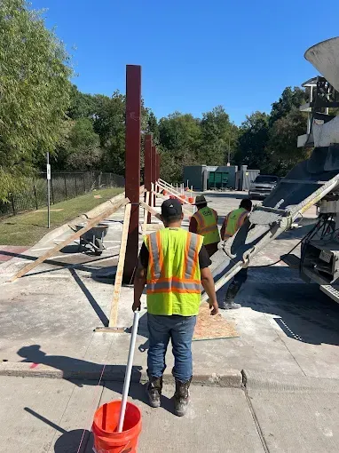 Workers in high-visibility vests pour concrete near vertical steel beams at an outdoor construction site.