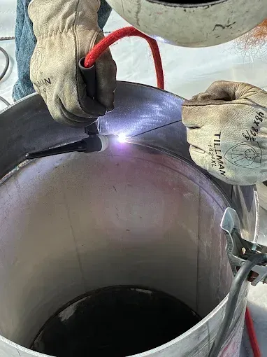 A welder wearing gloves performs TIG welding on a cylindrical metal pipe, creating a bright white arc at the seam.